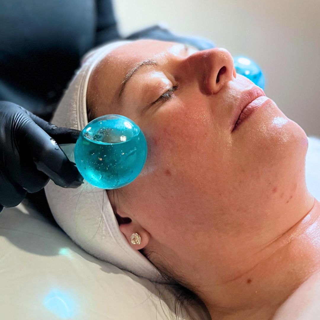 Woman receiving a facial treatment with blue, liquid-filled cryo globes held by black-gloved hands.