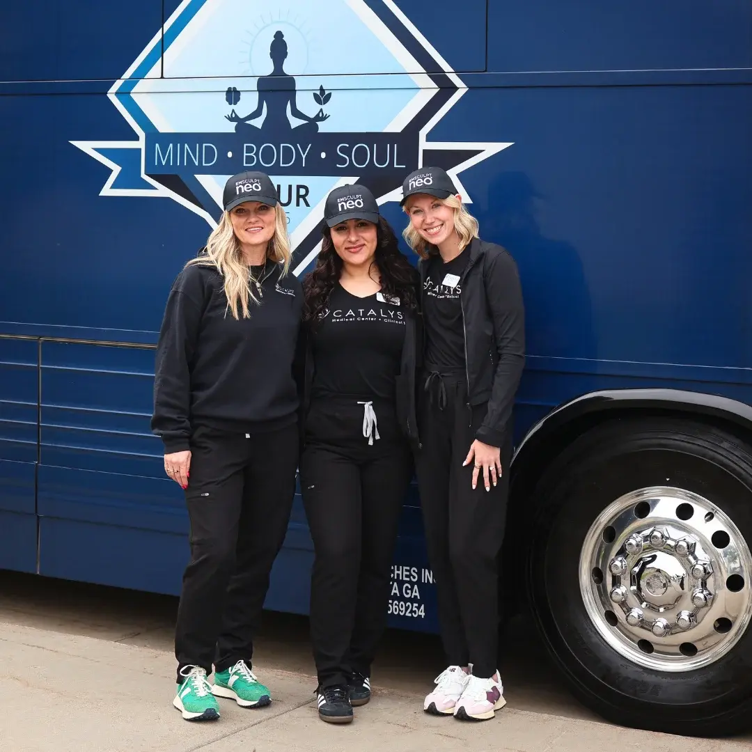 Happy coworkers with black Catalyst Medical Center + Clinical Spa shirts and Emsculpt neo baseball caps pose for a photo in front of a blue tour bus.