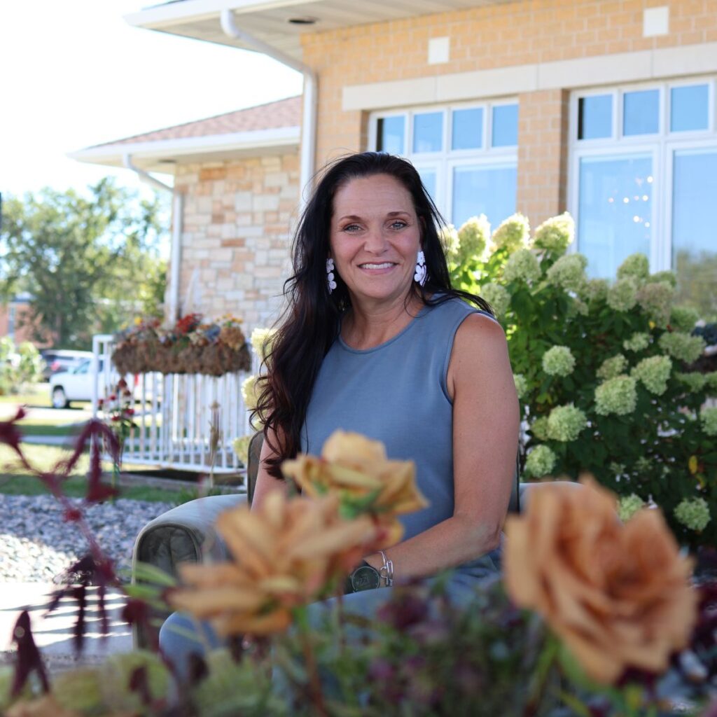 Woman with dark hair in a blue top sits smiling at the camera outside a medical facility with flowers all around.