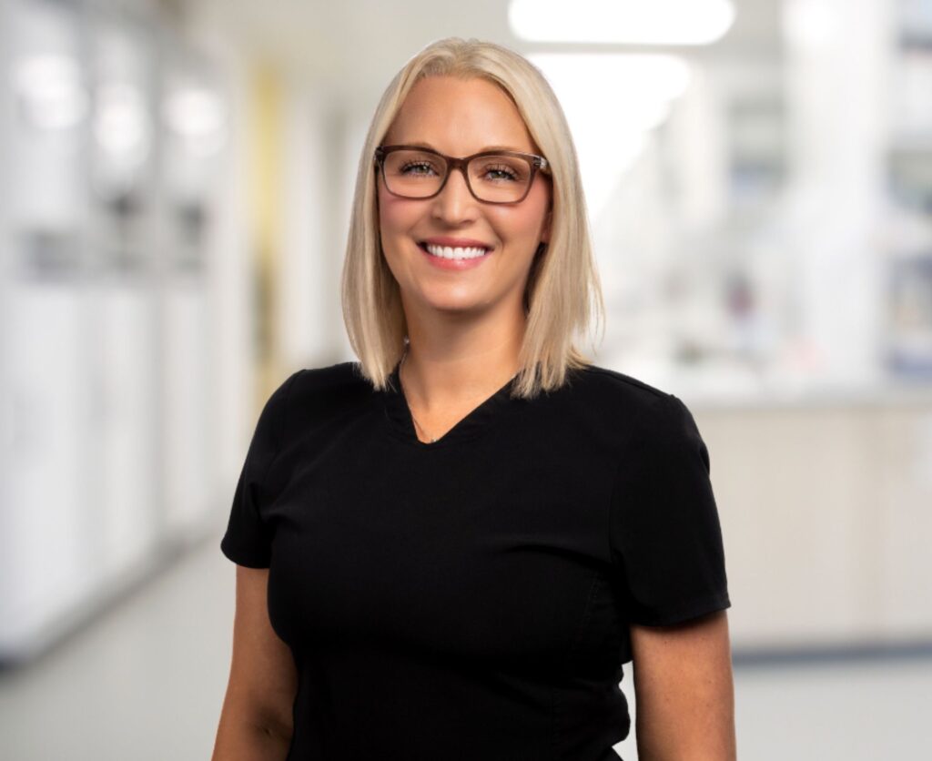 Blonde woman with glasses and a black shirt smiles at the camera in a clinic setting.